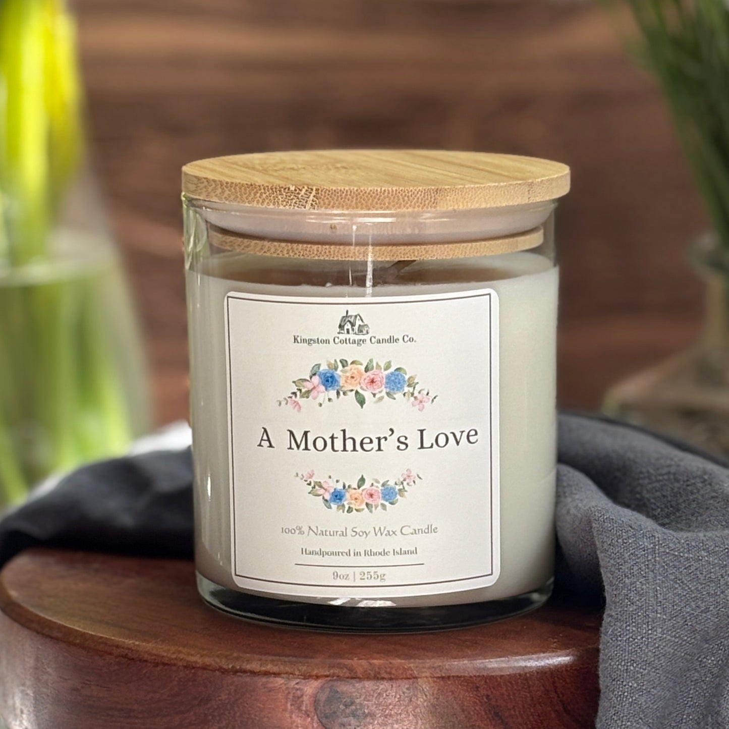 Candle jar labeled 'A Mother's Love' on a wooden surface with flowers in the background