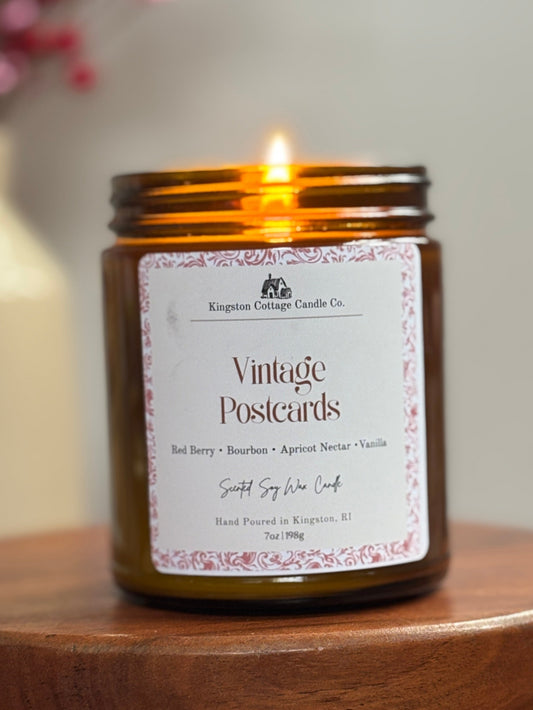 Candle in a glass jar labeled 'Vintage Postcards' on a wooden stand with a blurred background