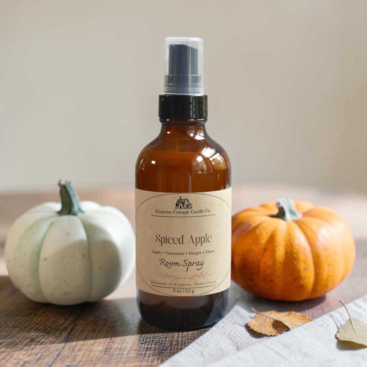An amber glass room spray bottle with a cream label reading 'Spiced Apple' by Kingswell Cottage Candle Co., styled on a wooden surface with a small white pumpkin on the left and a small orange pumpkin on the right.